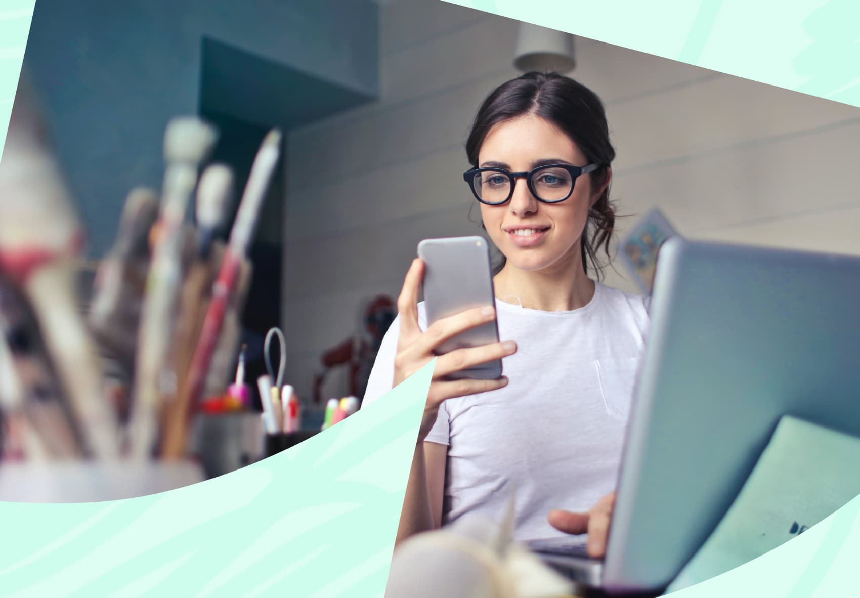 Woman looking at her mobile phone with a laptop on the desk in front of her