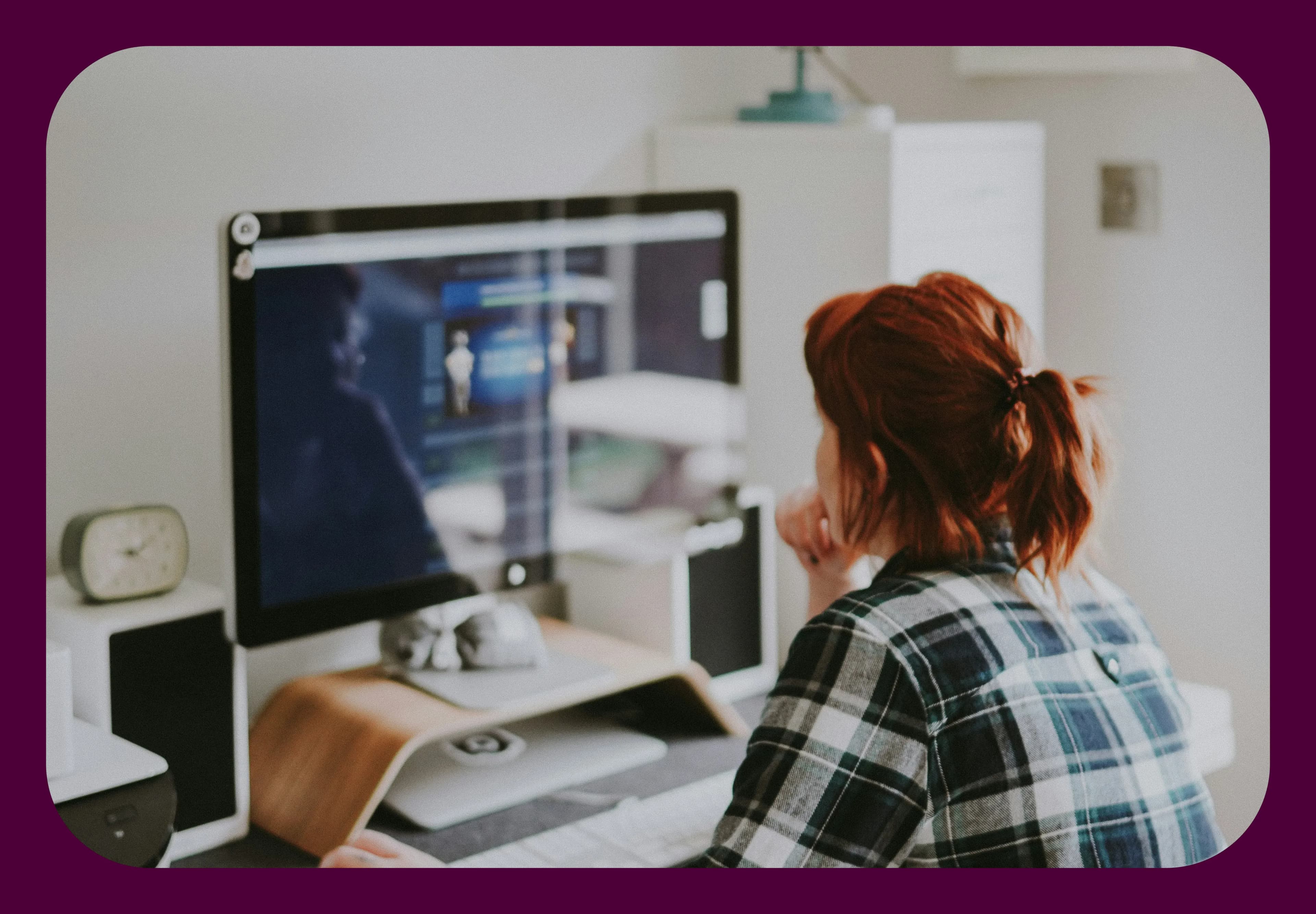 Person with red hair working on a computer at a desk, wearing a plaid shirt, in a home office setting with a clock and speakers nearby.